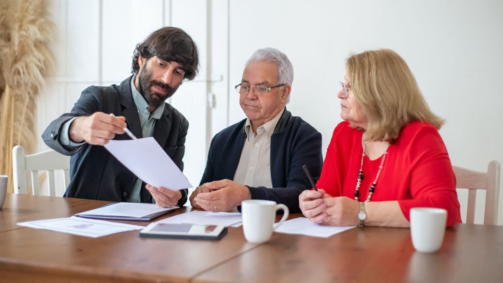 A man showing papers to an older couple seated at a wooden table with coffee mugs, a tablet and documents.