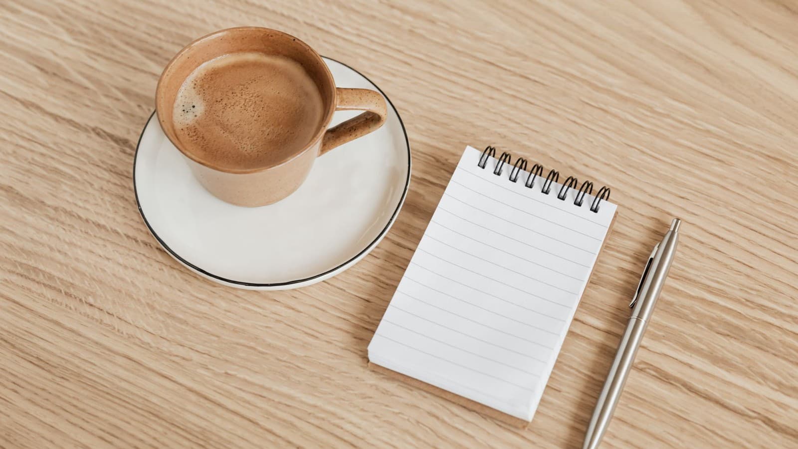 A cup of coffee on a saucer beside a blank spiral notepad and a pen on a wooden table.