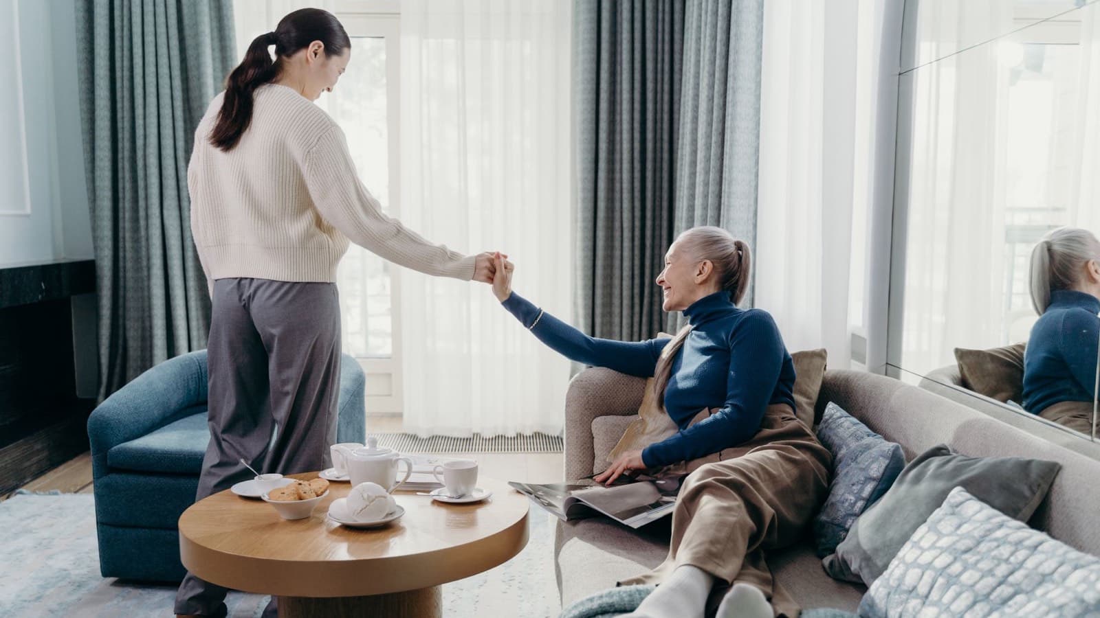 A smiling older woman on a sofa holding the hand of a younger woman standing beside her, with a tea set on the table.