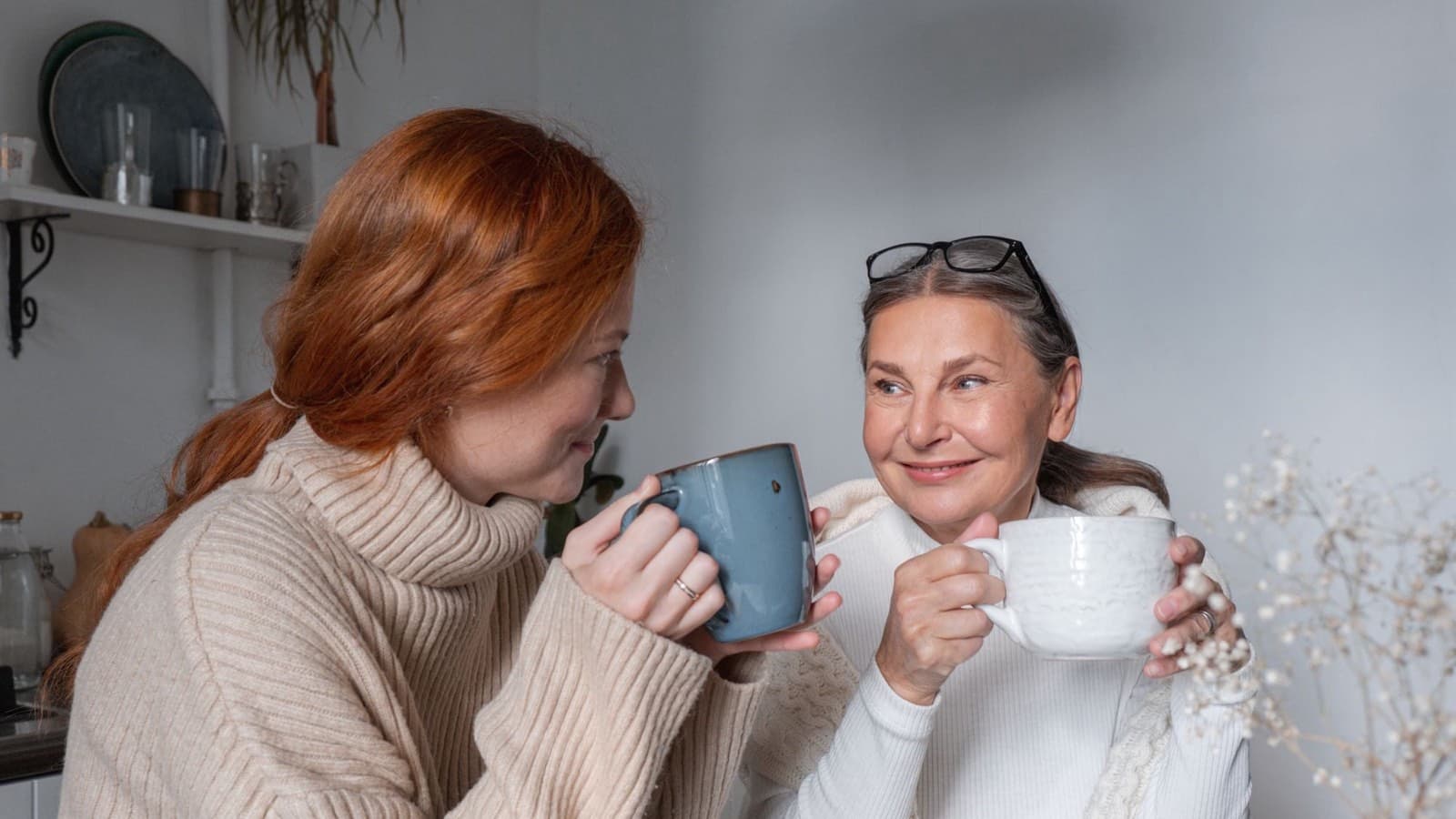 Two women sitting at a kitchen table, smiling at each other over mugs of coffee.