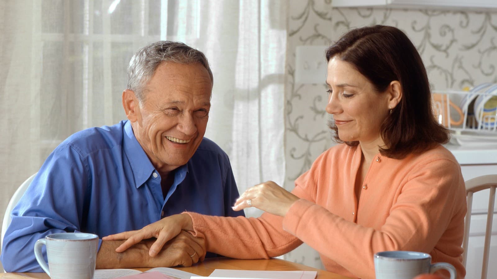 An older man laughing at a kitchen table while a woman rests her hand on his arm, with coffee mugs and papers in front of them.